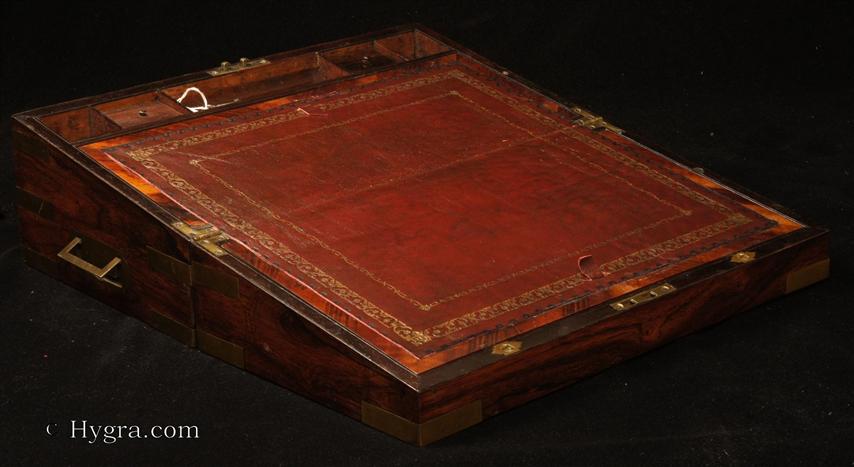 Figured rosewood writing box with brass corners,  straps, and  countersunk carrying handles having a side drawer and canceled secret drawers. The writing surface is framed by a cross -banding of rosewood and has been recovered with an embossed leather skiver.  The escutcheons both in front of the lock and the top are of ornate shape with spear like  fleur dis lis motif  to the side  The box has a working lock and key. Circa 1840.   Enlarge Picture